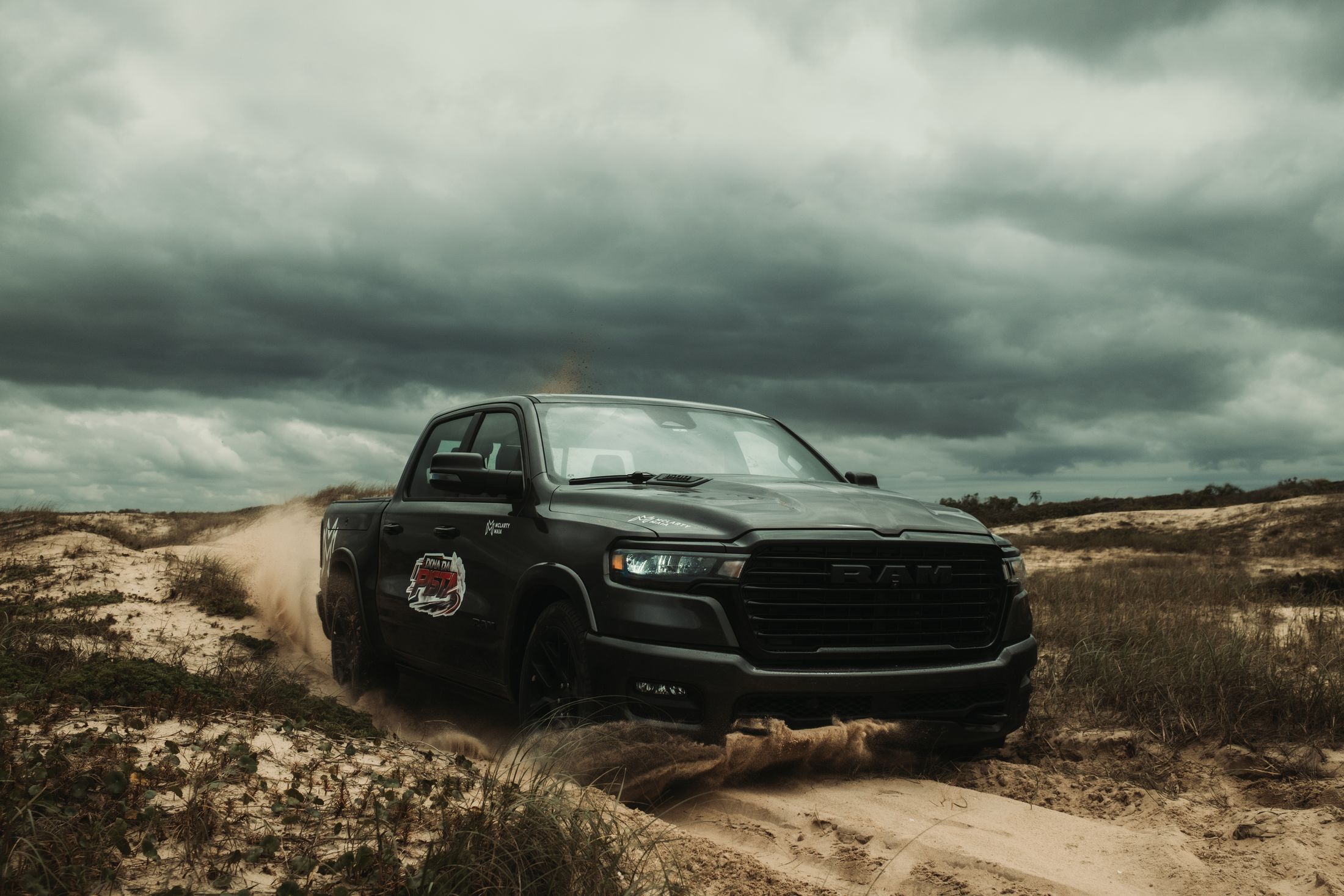 A black pickup with the Dona da Pista decal kicking up sand in the dunes under a storm sky
